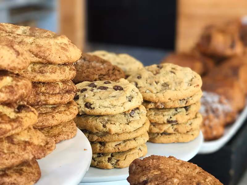Stacks of freshly baked cookies including snickerdoodle and chocolate chip varieties displayed on white platters at a bakery