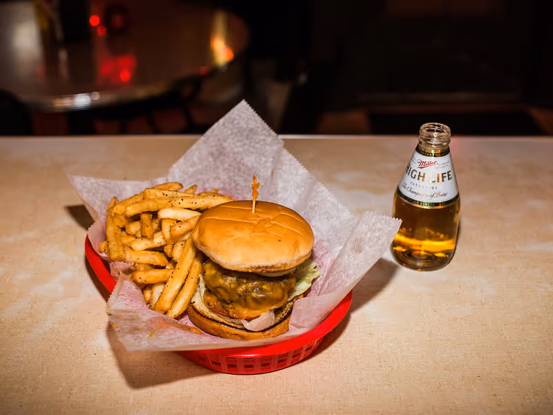 Cheeseburger and seasoned fries in a red basket served alongside a Miller High Life beer bottle on a restaurant table