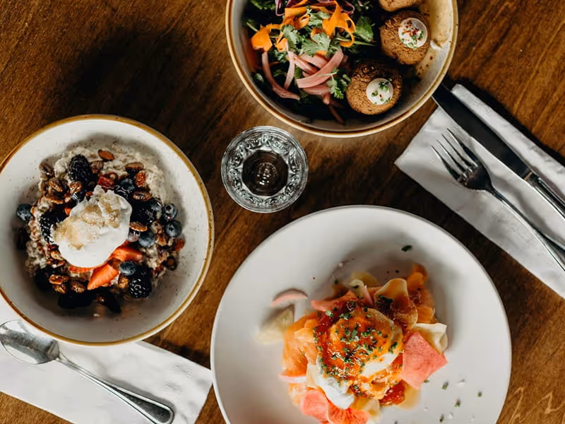 Overhead view of brunch dishes on a wooden table featuring berry oatmeal, a falafel salad bowl, and a smoked salmon plate