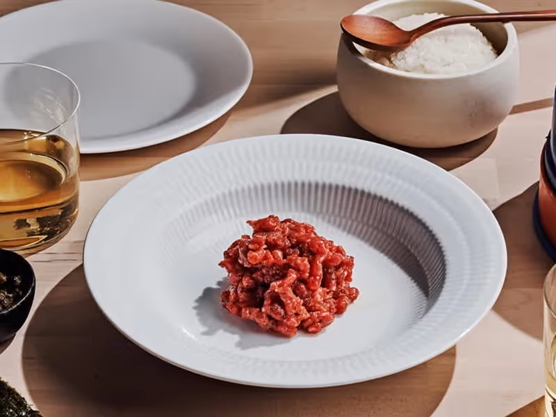 Raw beef tartare served on a white ribbed plate alongside steamed rice in a ceramic bowl and a glass of white wine.