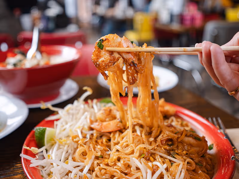 Chopsticks lifting shrimp and rice noodles from a red plate of Pad Thai garnished with bean sprouts and lime wedges