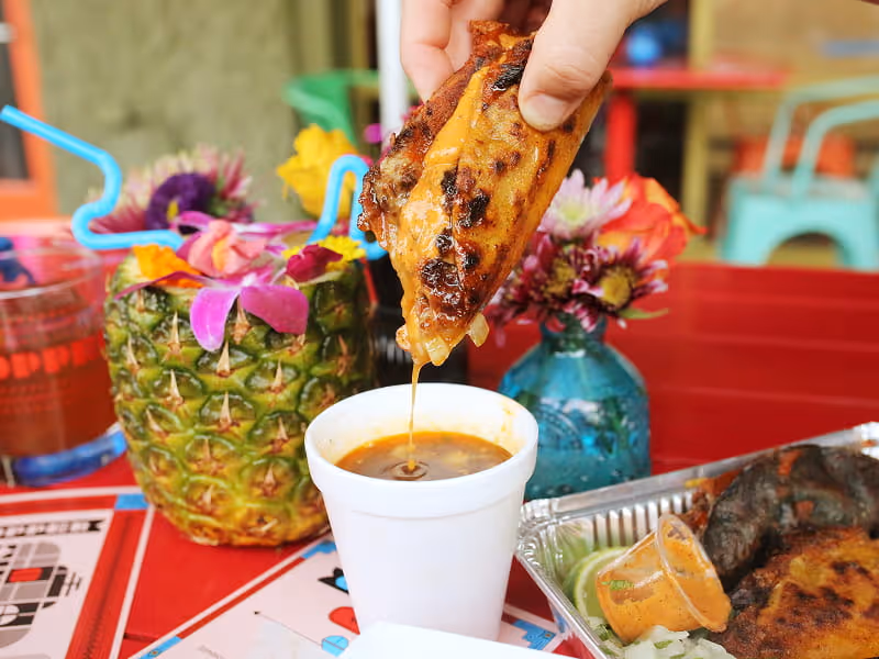 Hand dipping a grilled birria taco into a cup of rich consommé at a colorful tropical-themed outdoor restaurant.