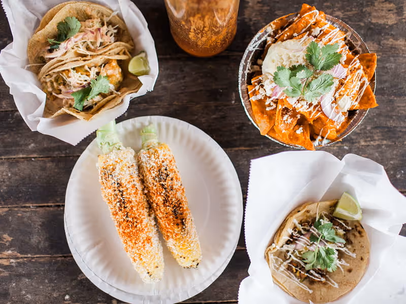 Mexican street food spread featuring elote corn on the cob, tacos with cilantro, and chilaquiles on a rustic wooden table.