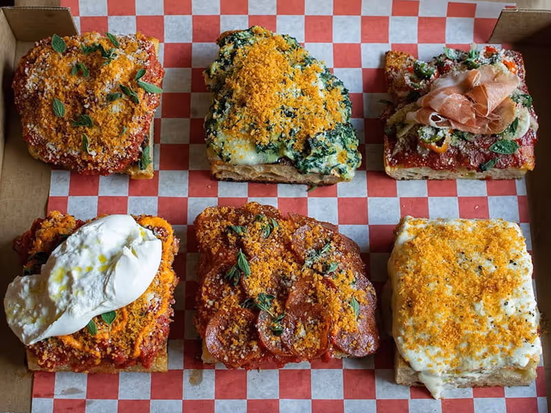 Six square slices of Detroit-style pizza with various toppings including pepperoni, spinach, burrata, and prosciutto