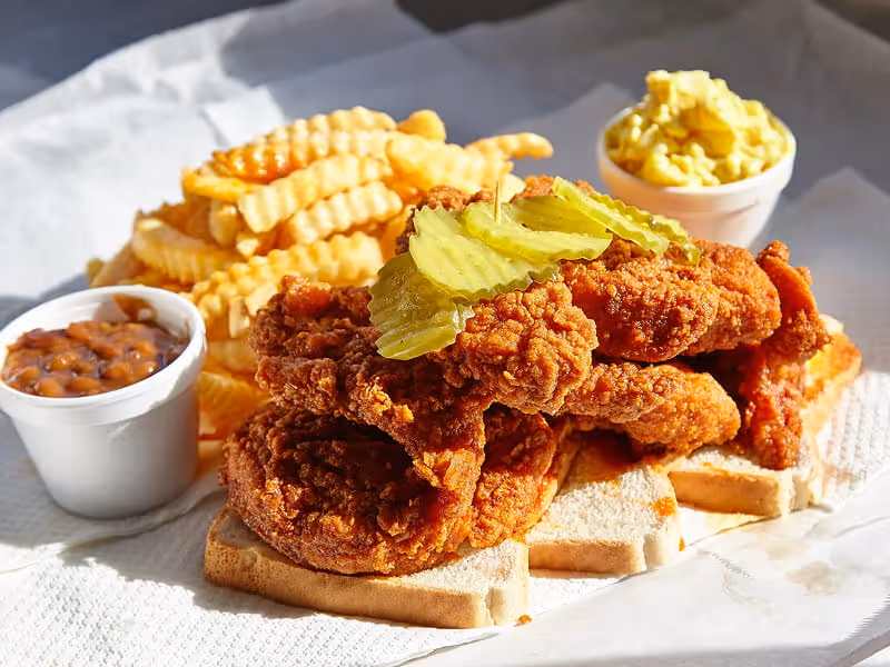 Nashville hot chicken tenders on white bread with crinkle-cut fries, pickles, baked beans, and potato salad on the side.