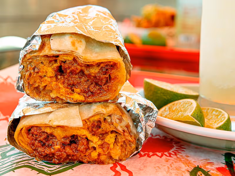 A burrito cut in half and stacked, wrapped in foil, showing a meaty filling, served with fresh lime wedges on the side.