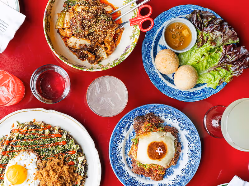 A colorful spread of Korean-inspired dishes on a red table, featuring rice balls, lettuce wraps, stir-fry, and fried egg