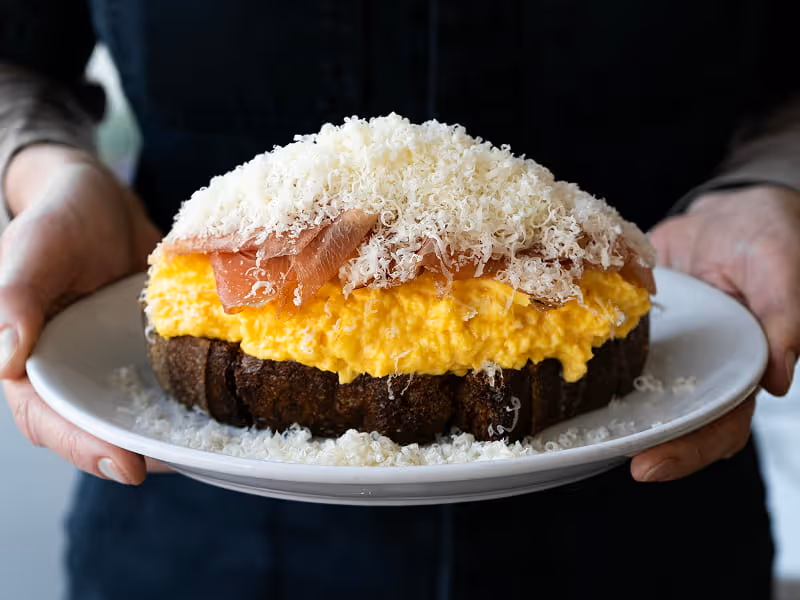 A person holds a plate of scrambled eggs on dark bread topped with prosciutto and finely grated Parmesan cheese.