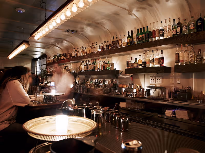 Intimate speakeasy-style cocktail bar with arched ceiling, backlit liquor shelves, and bartender preparing drinks in dim