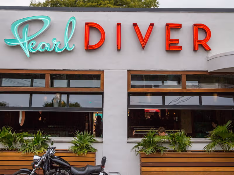 Pearl Diver restaurant exterior featuring retro neon signage in teal and red, tropical plants, and a motorcycle parked out