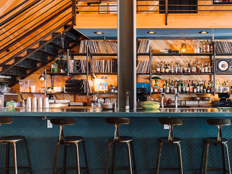 Retro bar interior with teal herringbone paneling, dark metal stools, vinyl record collection, and wooden staircase above.
