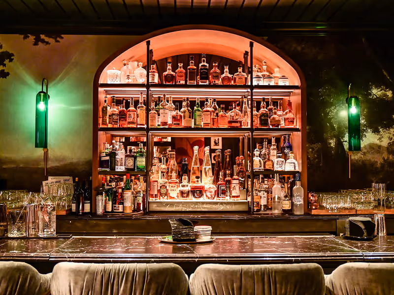 Upscale cocktail lounge featuring an arched backlit liquor display, dark marble bar top, velvet seating, and moody green