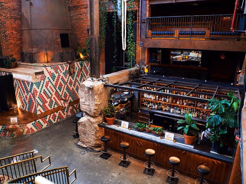 Industrial-style bar interior with exposed brick walls, colorful tile mosaic, well-stocked shelves, and leather bar stools