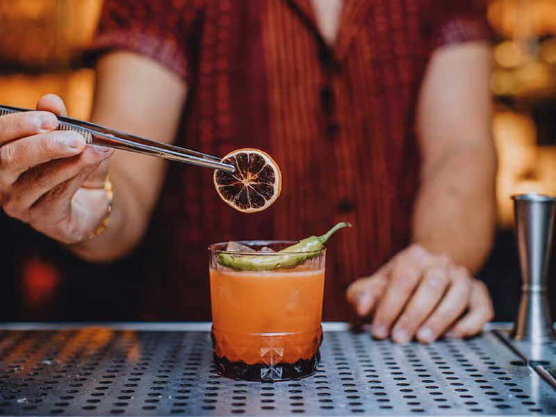 Bartender placing a dehydrated citrus wheel garnish on an orange cocktail with a chili pepper using metal tongs.