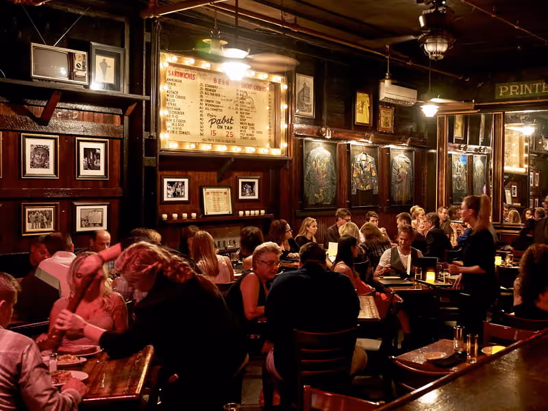 Crowded vintage bar interior with illuminated menu board advertising Pabst beer, framed photos on dark wood walls