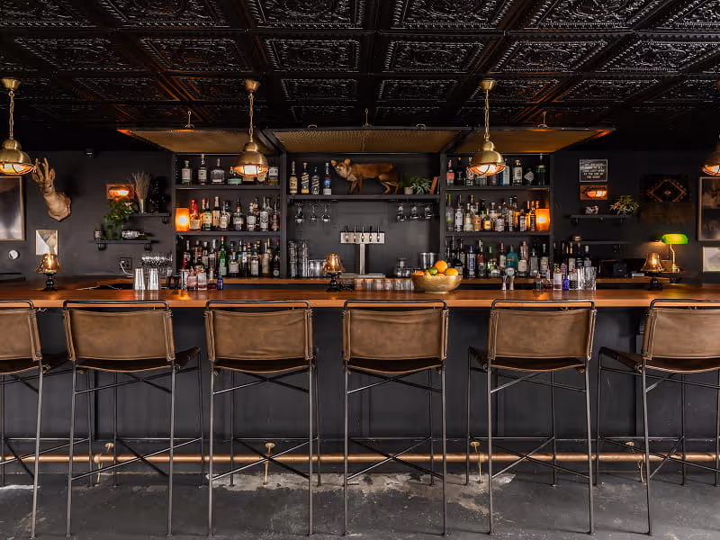 Moody speakeasy-style cocktail bar with dark tin ceiling, leather bar stools, brass pendant lights, and curated bottle