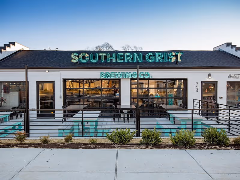 Southern Grist Brewing Co. exterior at dusk featuring teal neon signage, white brick facade, outdoor patio seating,