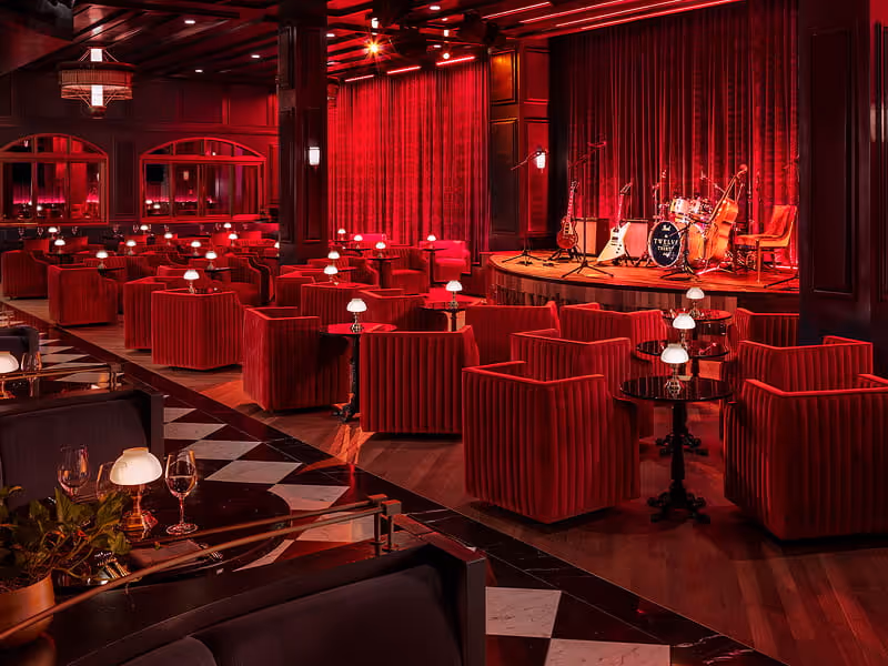 Upscale jazz lounge with red velvet armchairs, a small stage with drum kit and guitars, red curtains, and intimate table