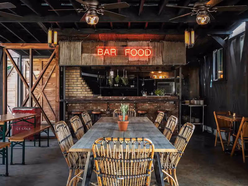 Rustic bar food counter with red neon BAR FOOD sign, corrugated metal accent, rattan chairs around a long wooden communal