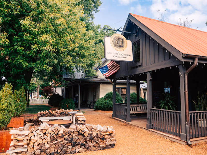 Leiper's Creek Gallery storefront with an American flag, rustic dark wood porch, copper roof, and stacked firewood out front.