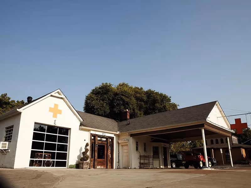 Converted vintage gas station with white exterior, yellow cross symbols, large garage door windows, and a covered carport