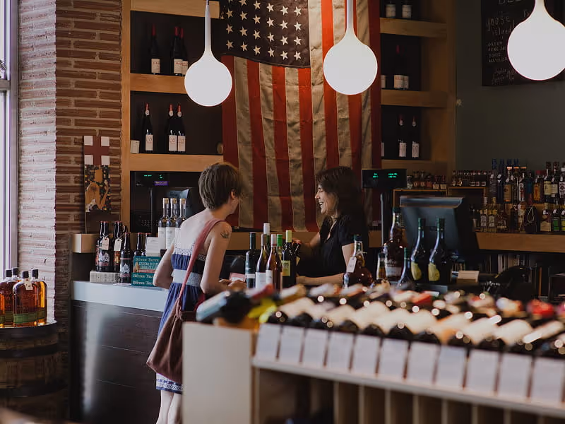 Two women chatting at a wine and spirits shop counter with a large American flag displayed on the wall behind them.