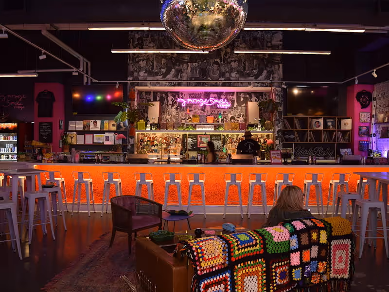 Retro bar interior with orange-lit counter, disco ball, neon signage, white stools, and a cozy couch with a crocheted