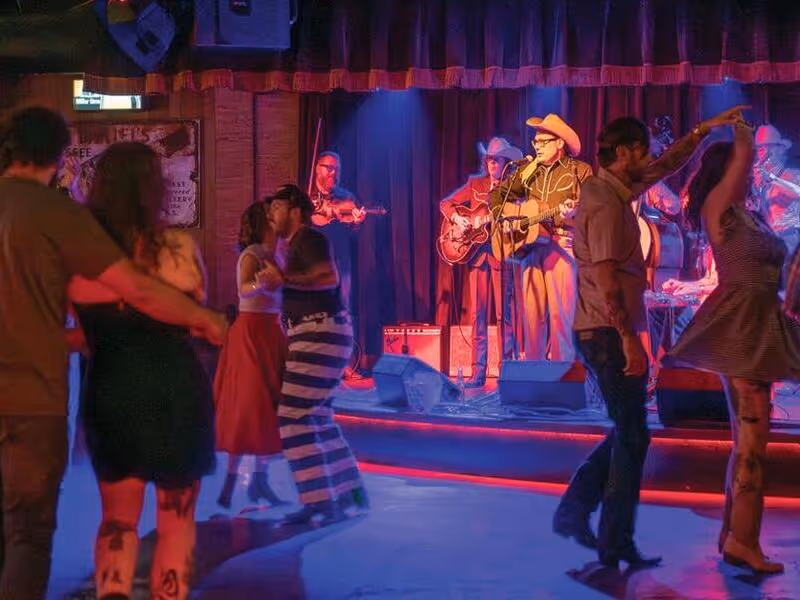 Couples dance at a honky-tonk bar while a country band with a cowboy-hatted guitarist performs on stage.