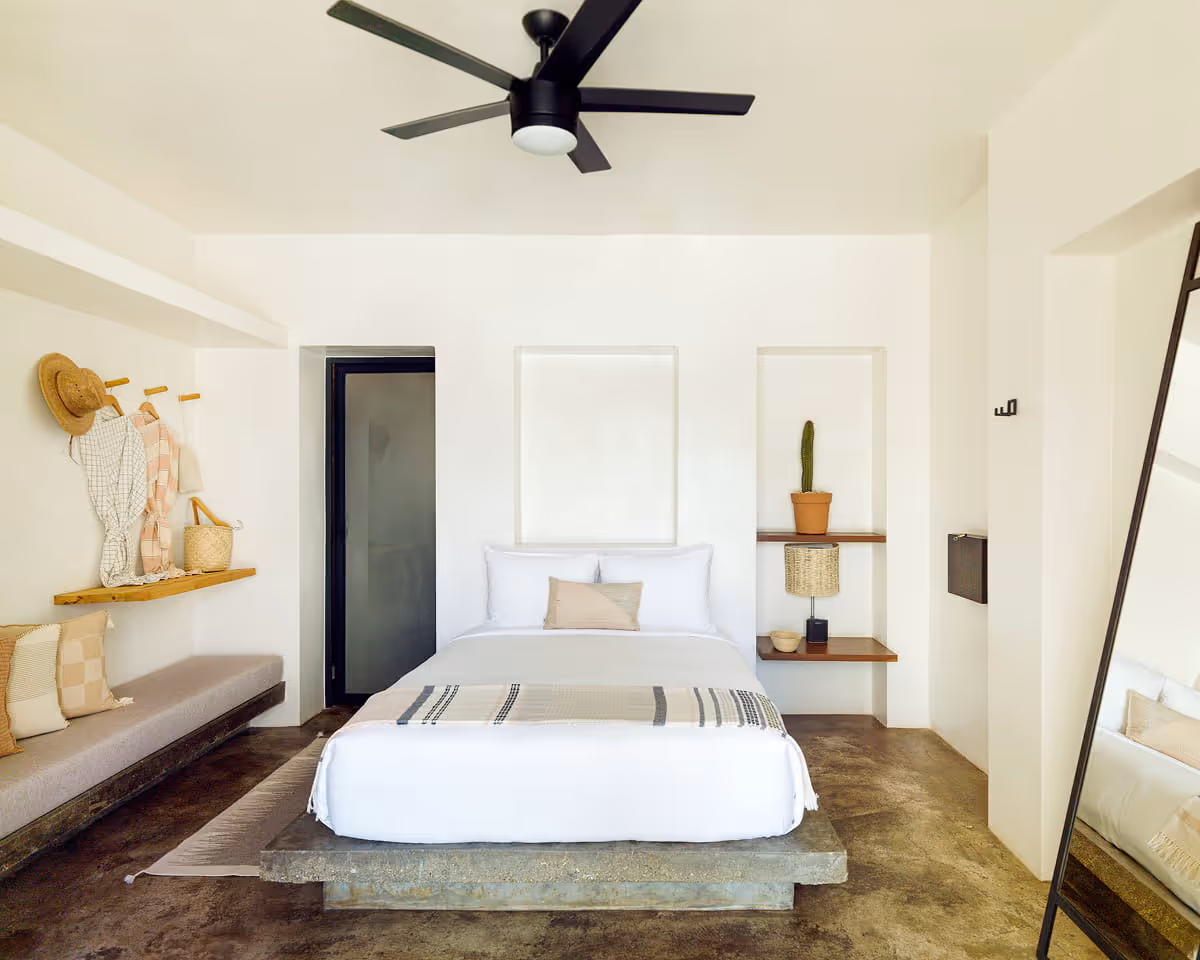 Boutique hotel bedroom featuring a concrete platform bed with white linens, black ceiling fan, wooden shelves, and a daybed