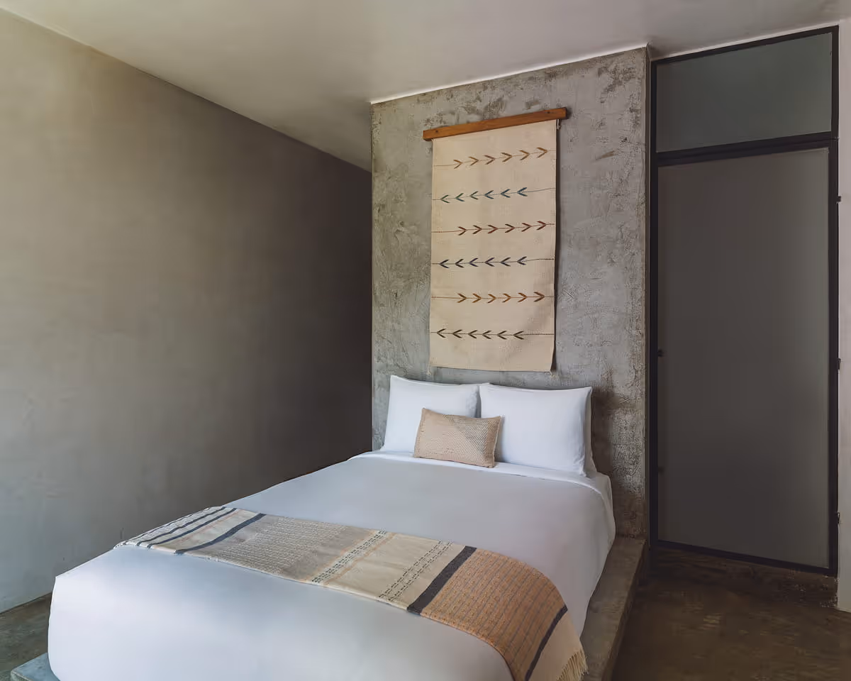 Cozy bedroom with raw concrete accent wall, white bedding, woven wall tapestry with arrow pattern, and polished concrete
