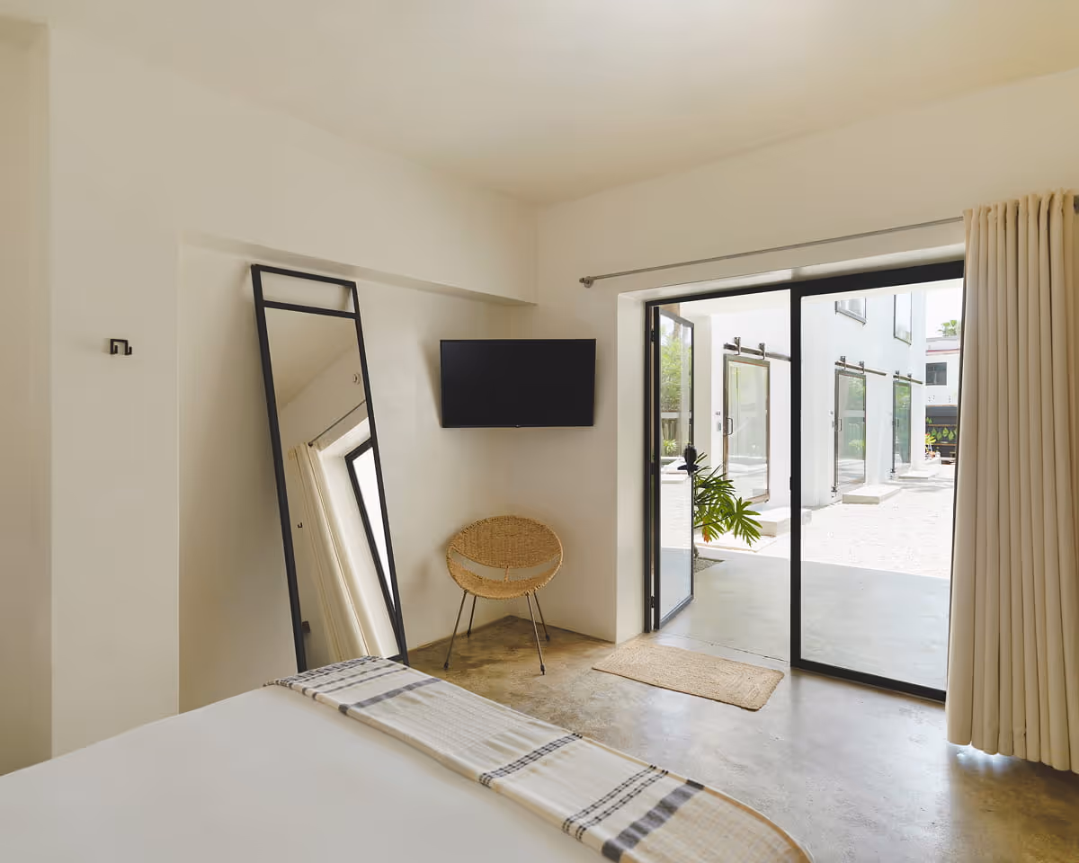 Minimalist bedroom with polished concrete floors, wall-mounted TV, wicker chair, and glass doors opening to a bright