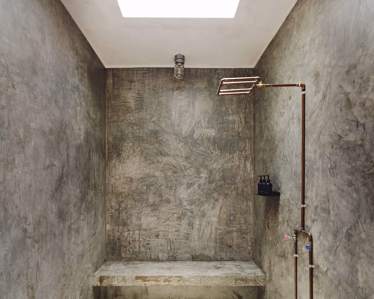 Industrial-style concrete shower with exposed copper plumbing, skylight overhead, built-in bench, and wall-mounted toiletry