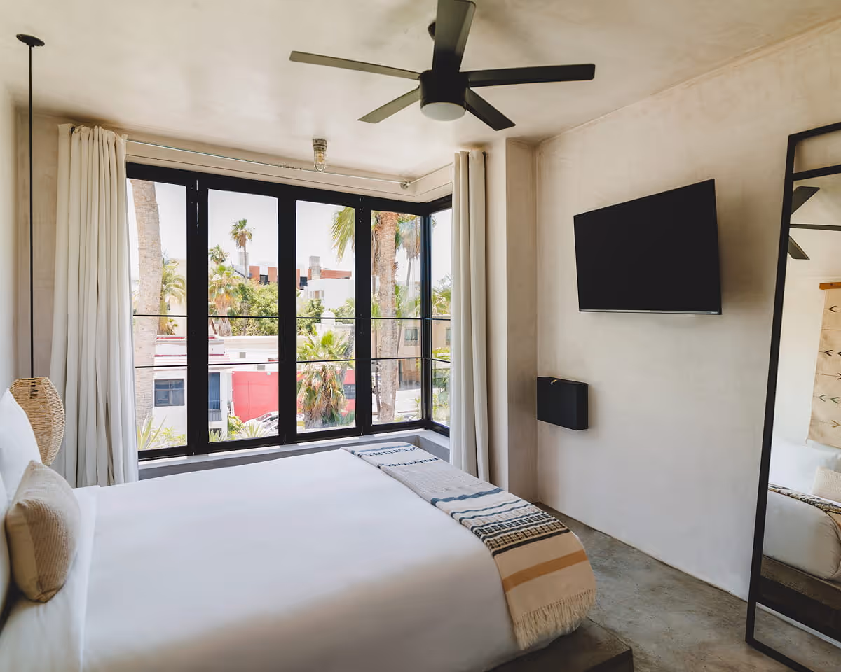 Minimalist hotel room featuring a white bed with striped throw blanket, wall-mounted TV, ceiling fan, and corner windows