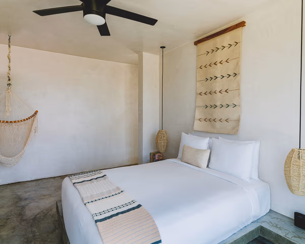 Cozy hotel bedroom with white bedding, woven arrow-pattern wall hanging, rattan pendant lights, hammock, and polished