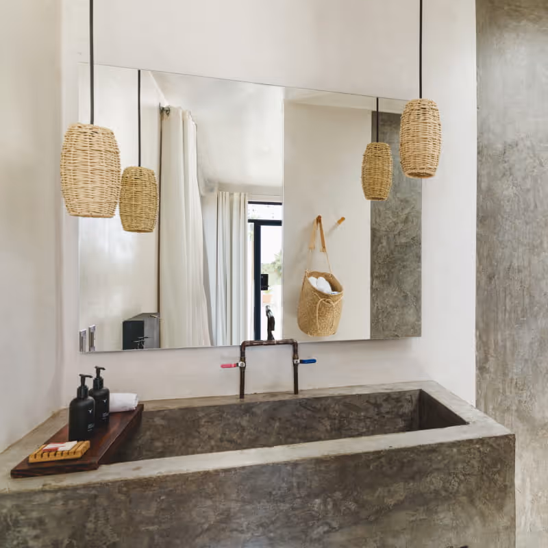 Concrete bathroom vanity with large mirror, woven rattan pendant lights, dark soap dispensers on a wooden tray,