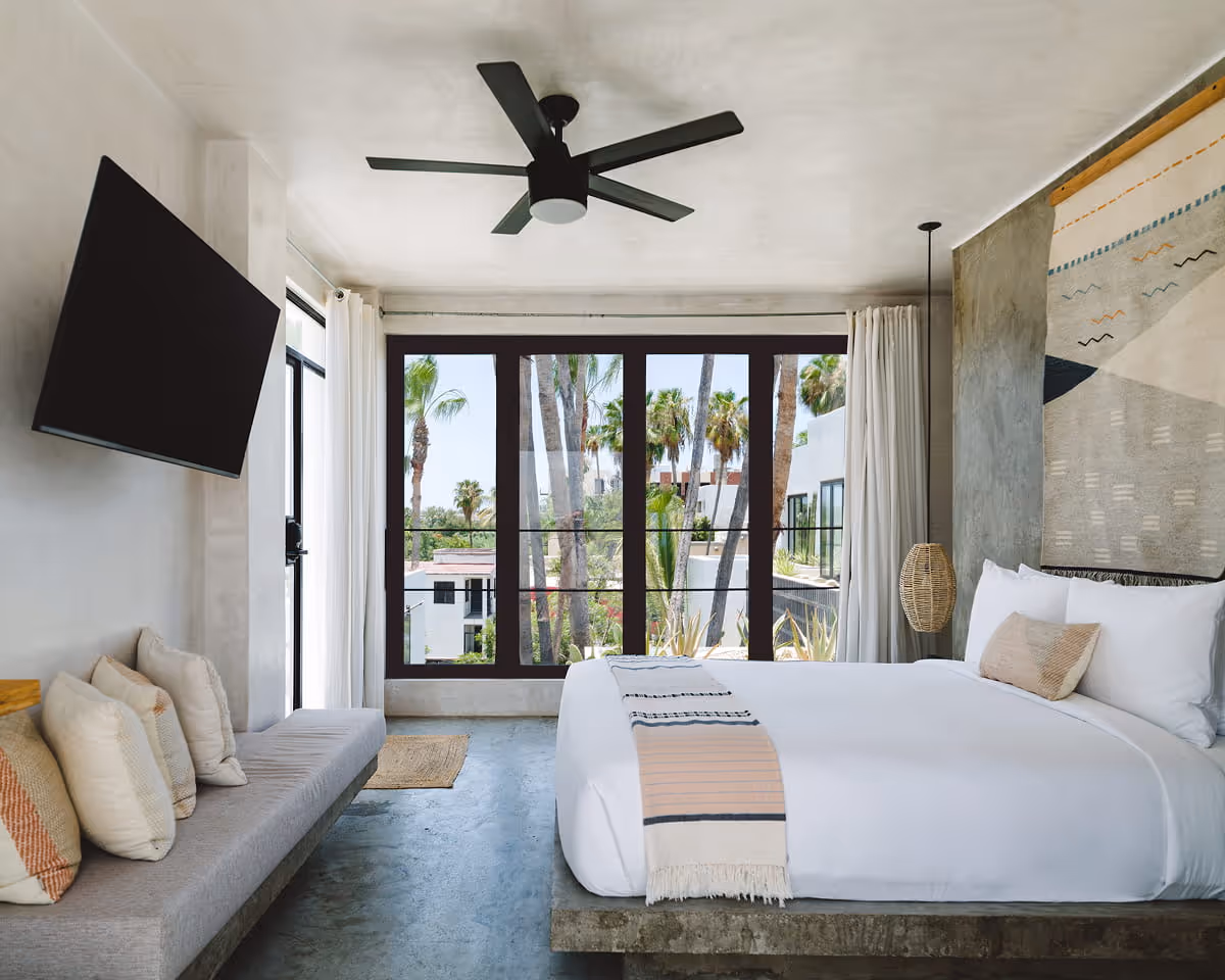 Boutique hotel bedroom with white bedding on a concrete platform, woven wall tapestry, daybed with pillows, and palm tree