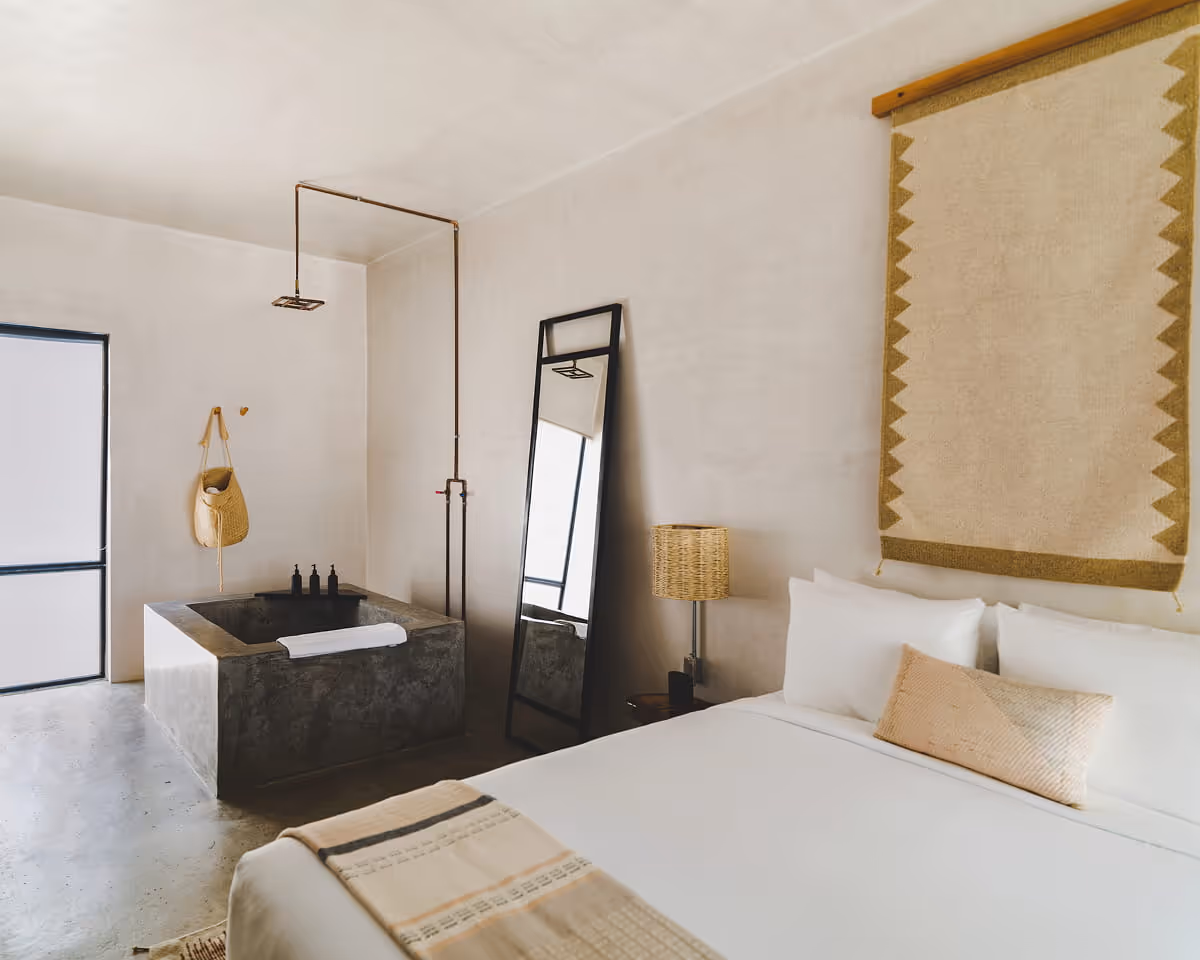 Rustic-chic hotel bedroom featuring a concrete soaking tub, copper rain shower, full-length mirror, and woven wall tapestry