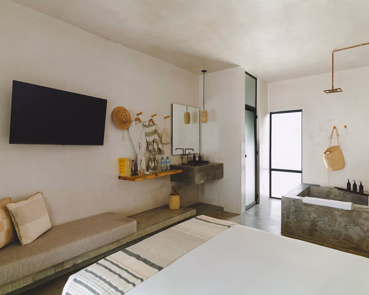 Open-concept hotel suite with wall-mounted TV, concrete daybed, floating wood shelf vanity, and freestanding concrete bathtub