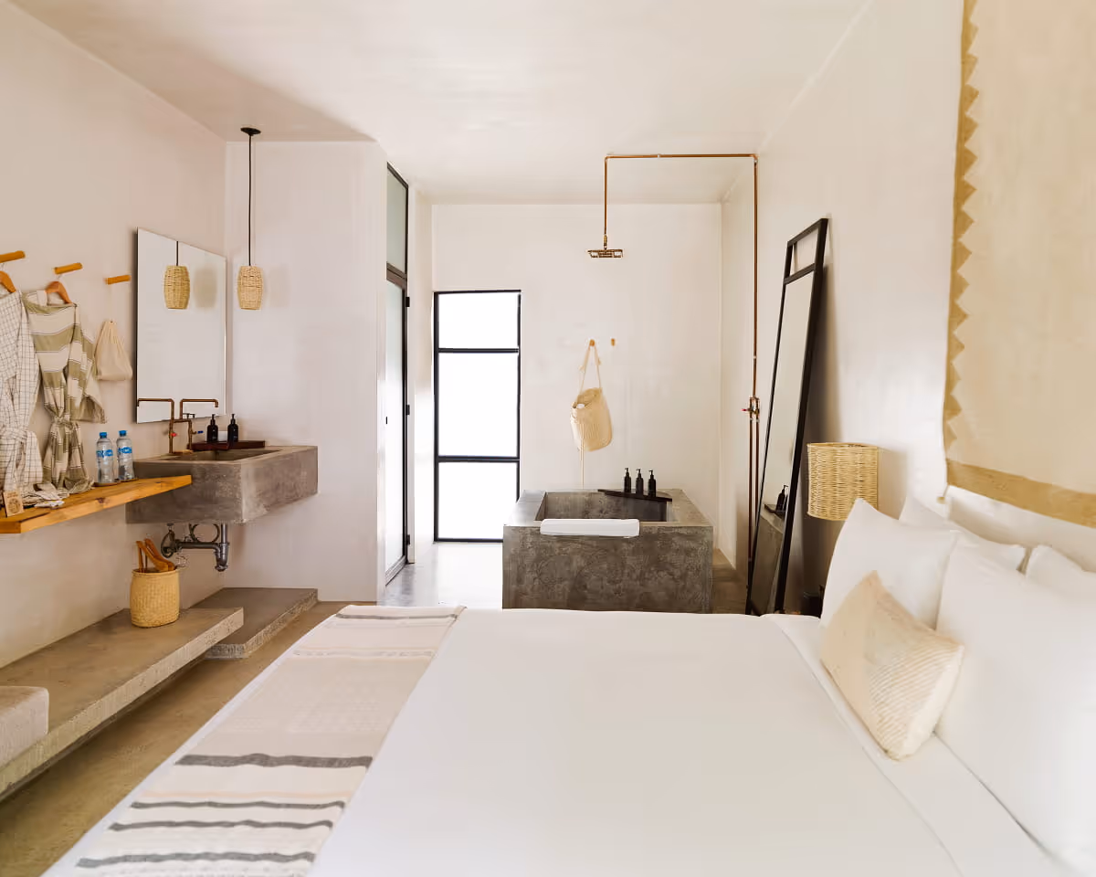 Minimalist boutique hotel room with white bed, concrete sink and bathtub, copper shower fixture, and woven basket accents