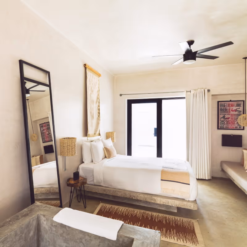Bright minimalist hotel room with white bedding, concrete platform bed, ceiling fan, full-length mirror, and Lucha Libre