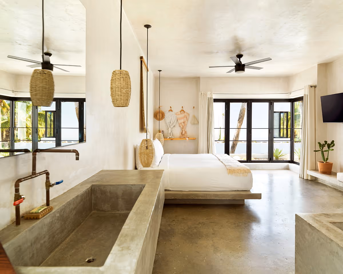 Open-concept suite featuring a concrete soaking tub, platform bed with woven pendants, exposed copper plumbing, and large