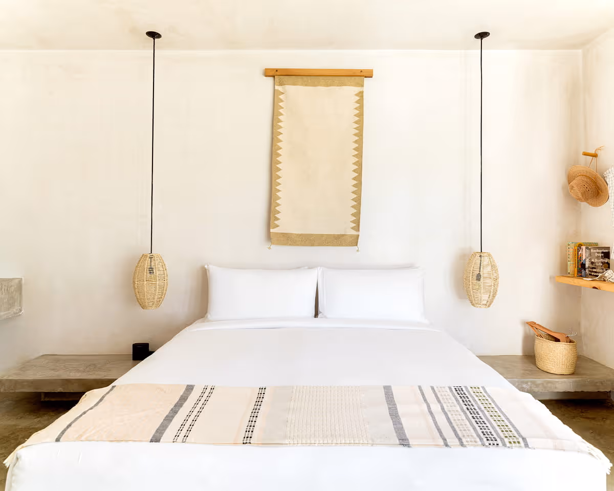 Close-up of a platform bed with crisp white linens, a woven throw blanket, rattan pendant lights, and a textile wall hanging