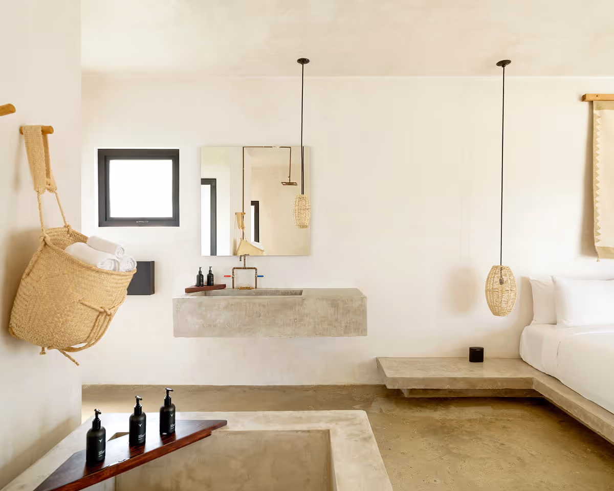Concrete floating vanity sink with mirror beside a platform bed, woven basket for towels, and artisan bath products