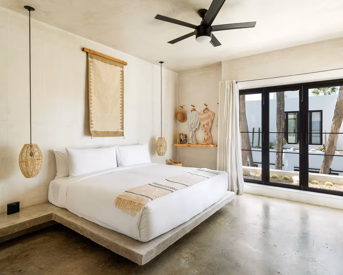 Minimalist hotel bedroom with concrete platform bed, woven pendant lights, textile wall hanging, and black-framed glass