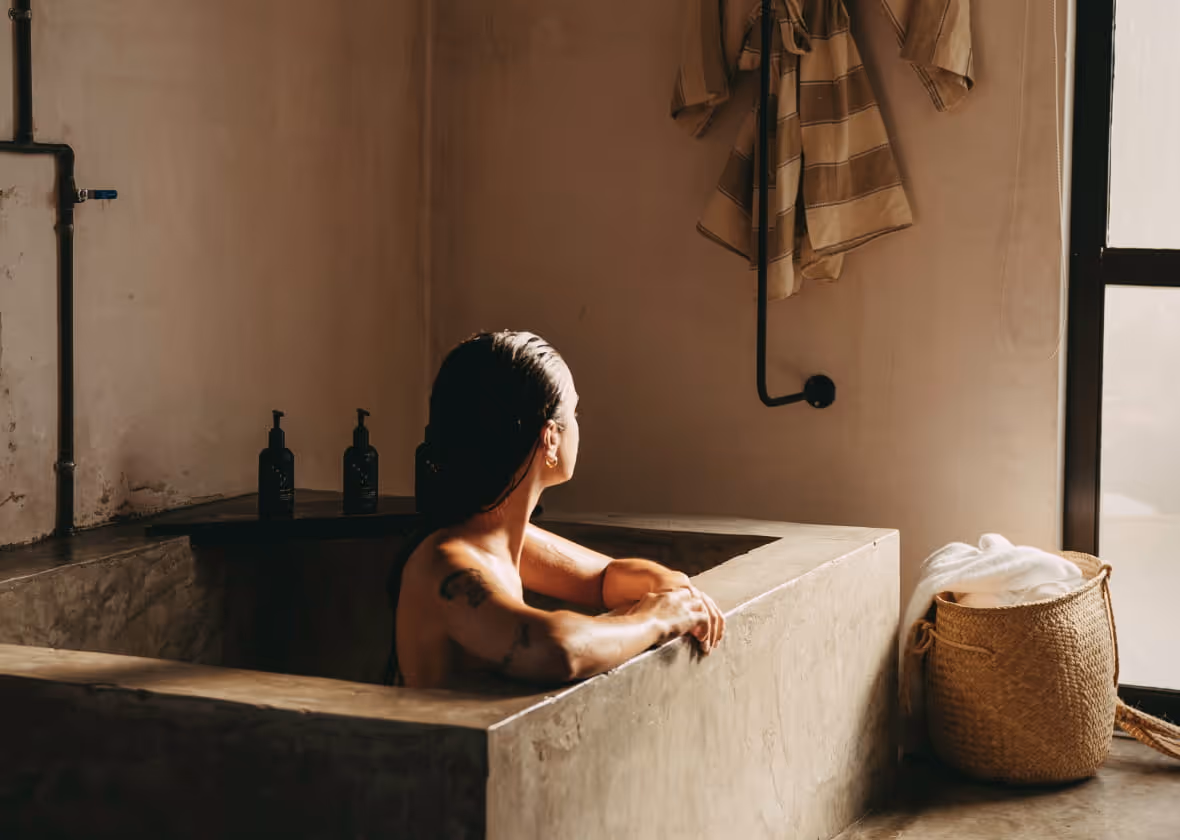 A woman relaxes in a concrete bathtub in a minimalist bathroom with warm earth tones and natural light.