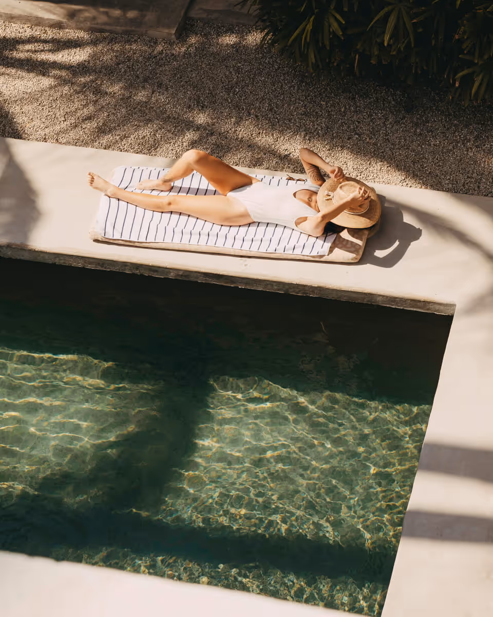 Woman in a white swimsuit lounging poolside on a striped towel with a straw hat covering her face in tropical sunlight