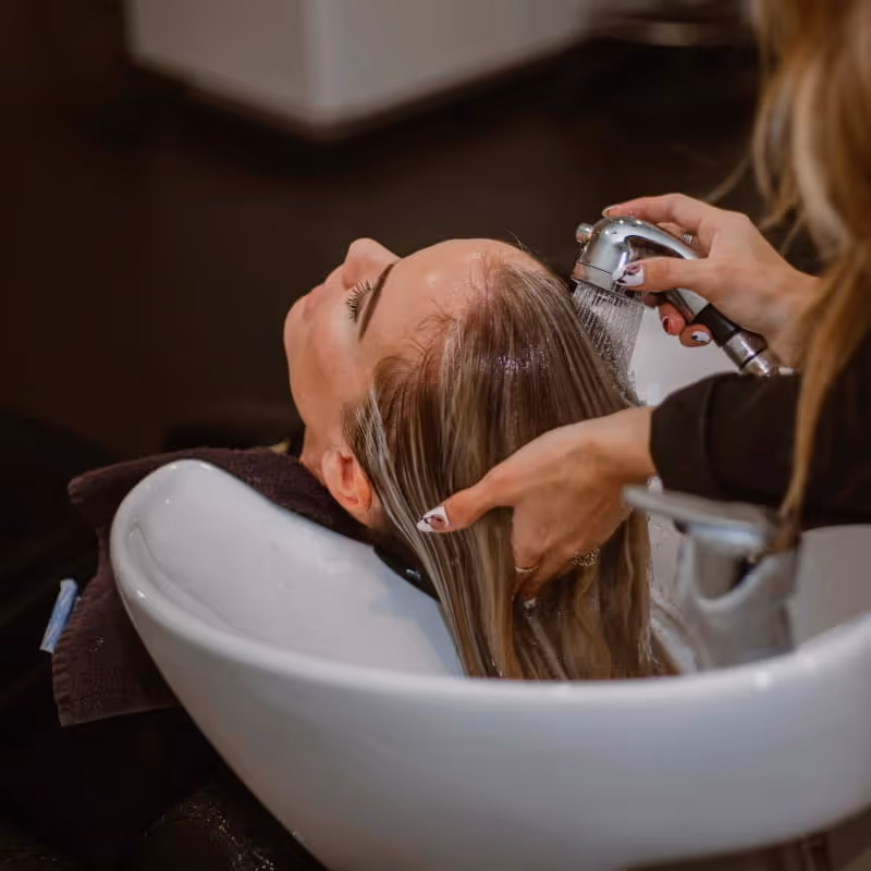 Hairstylist rinsing a client's blonde hair with a handheld sprayer over a white salon shampoo bowl during a wash treatment.