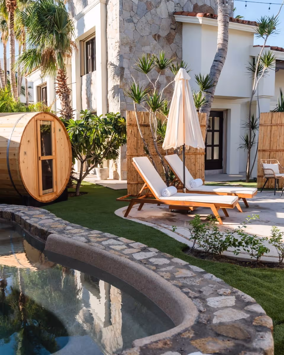 Luxury resort poolside area with wooden barrel sauna, white lounge chairs, umbrella, and lush tropical landscaping on green