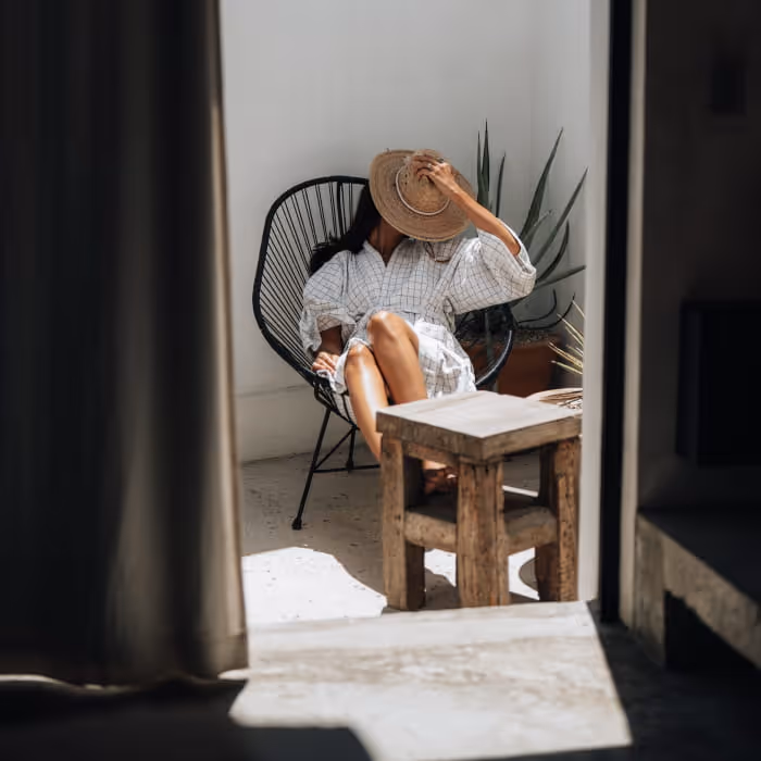 Woman holding a straw hat over her face while lounging in a black wire chair on a sunlit minimalist patio