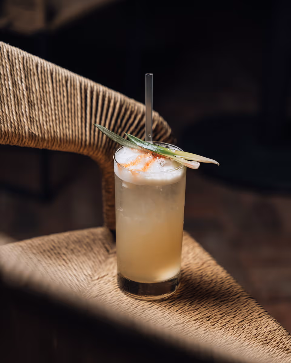 A tropical pineapple cocktail with a glass straw and pineapple leaf garnish resting on a woven rattan chair in warm light